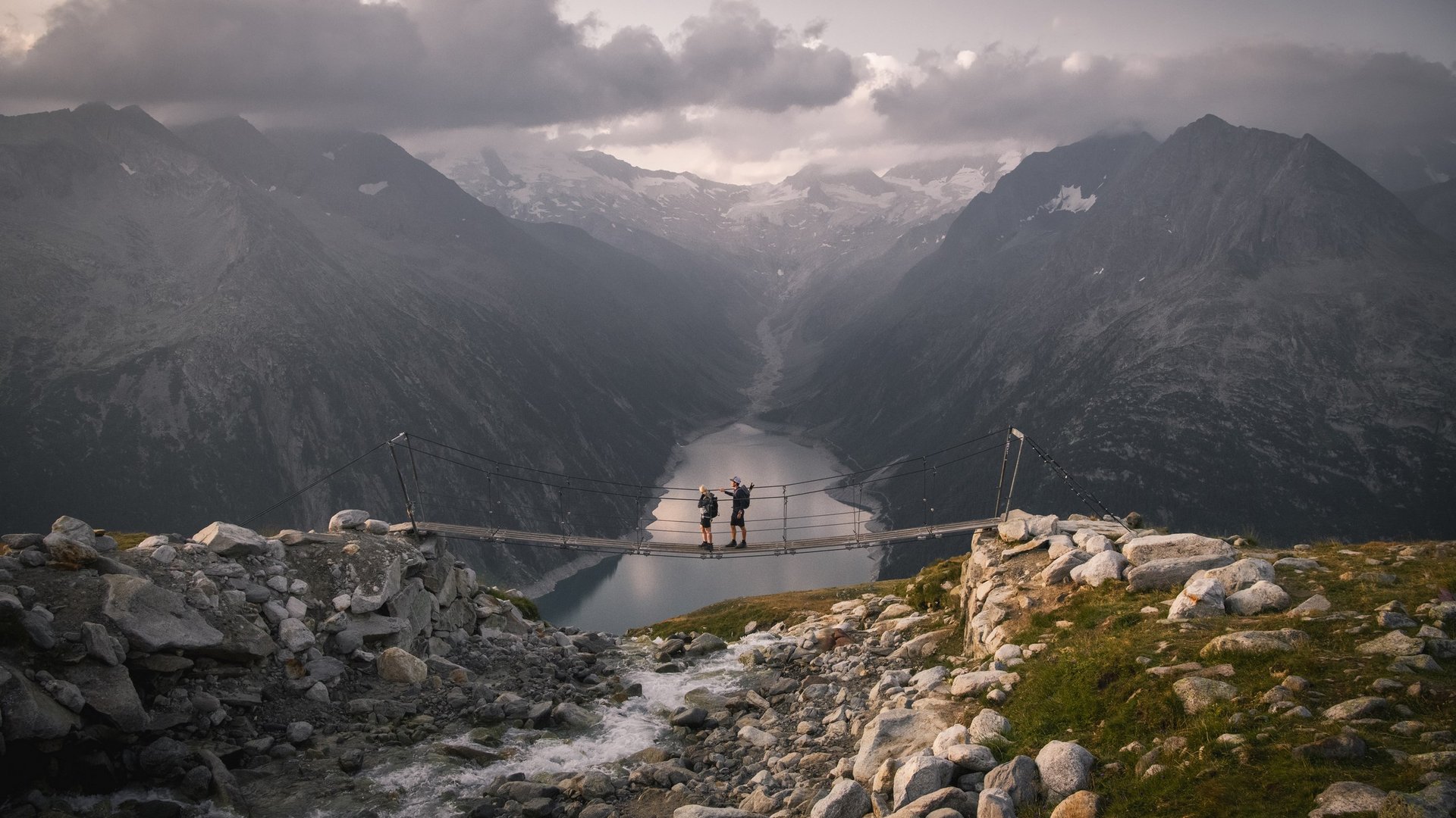 Hängebrücke über Bergsee Zwei Wanderer überqueren eine Hängebrücke mit Blick auf einen weitläufigen Bergsee und umliegende Gipfel.