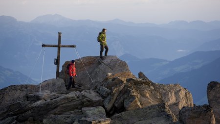Gipfelwanderung mit Kreuz Wanderer auf einem Gipfel neben einem großen Gipfelkreuz mit Panoramablick auf die umliegenden Berge.